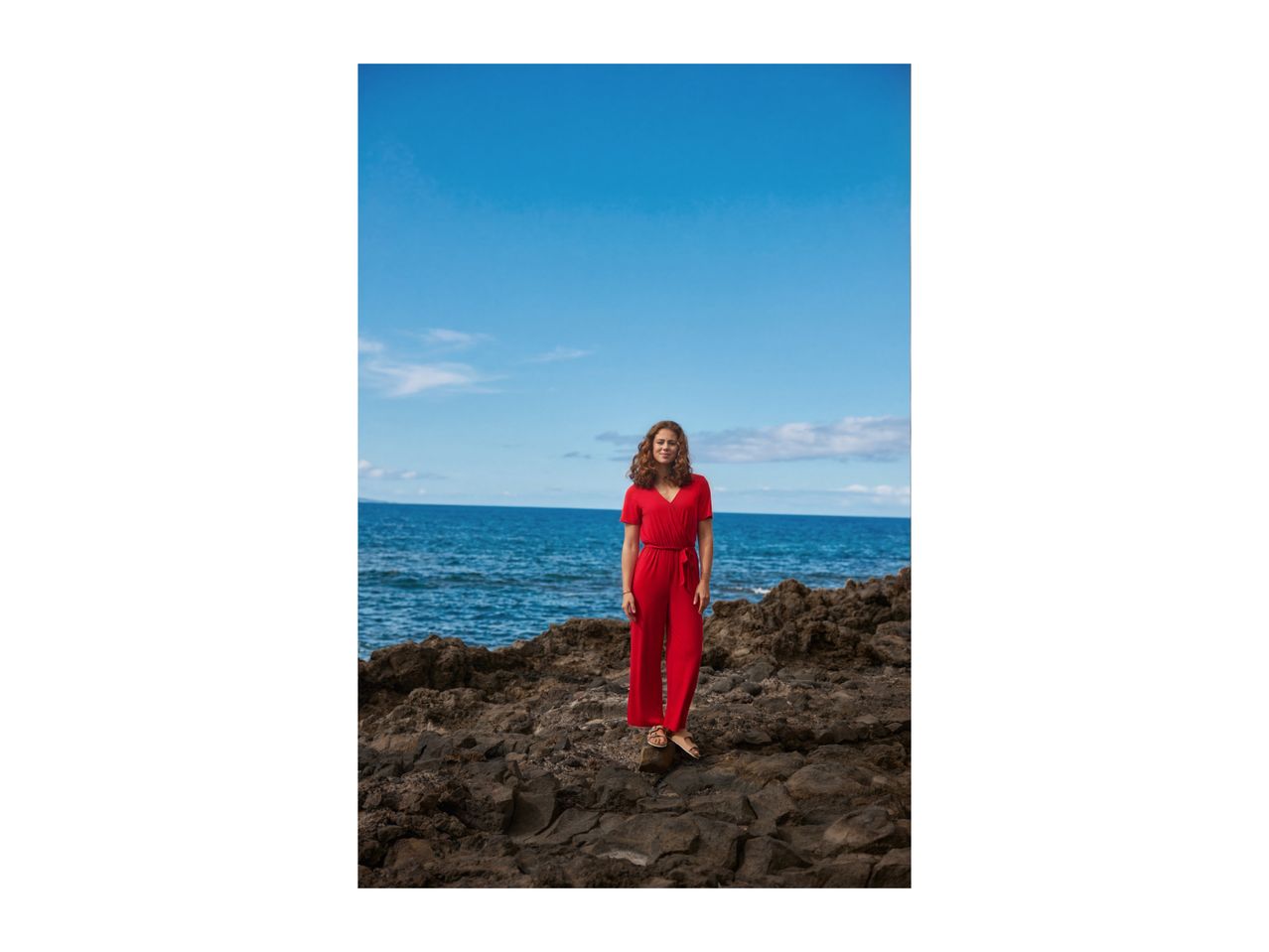 Woman in a red jumpsuit standing on rocks by the ocean under a blue sky.