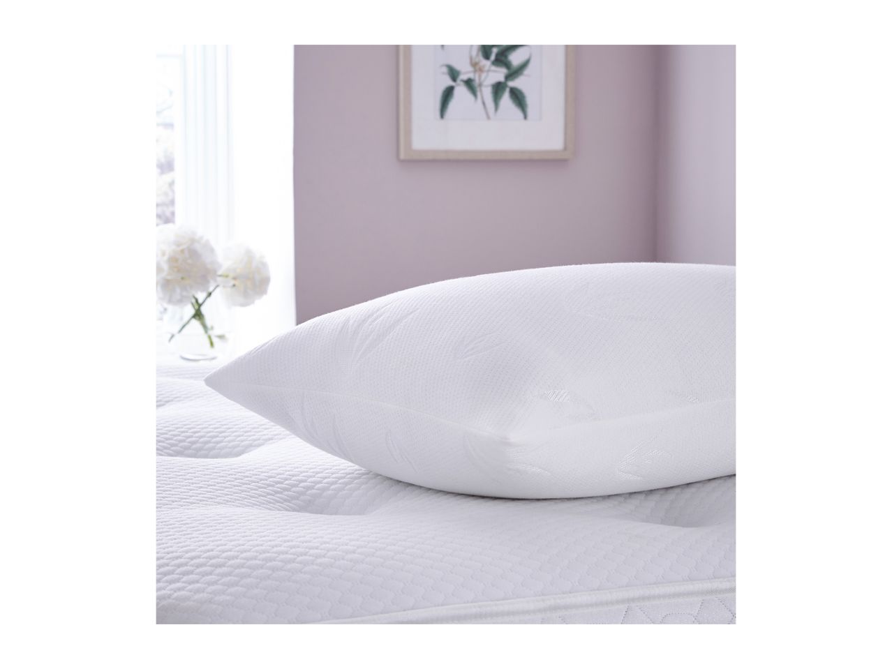A white pillow with a subtle pattern rests on a quilted mattress in a bright room.