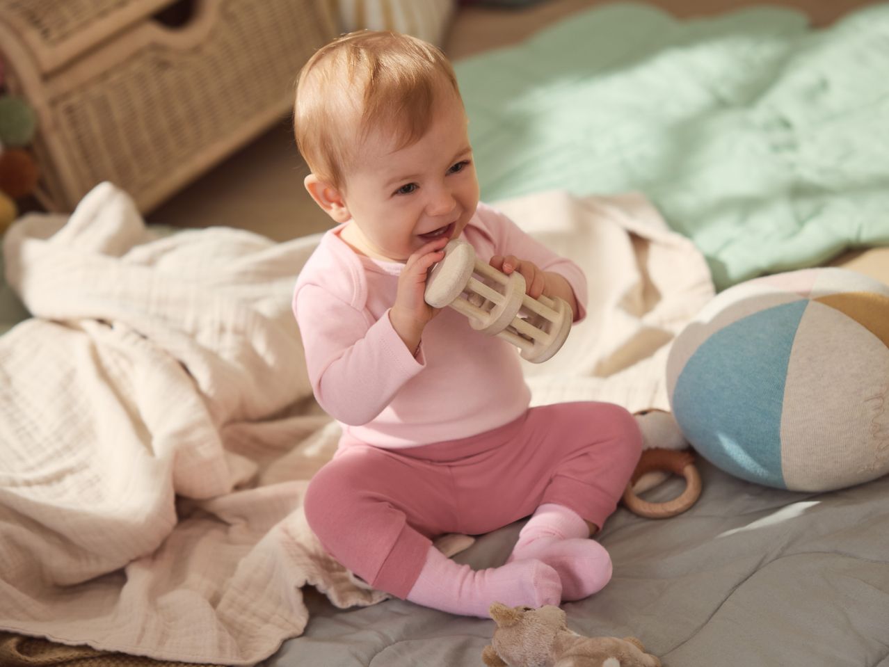 Happy baby in pink clothes playing with a wooden rattle on a soft blanket.