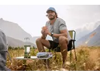 Man in cap and t-shirt sitting on a camping chair, holding a mug, with a lantern and table.