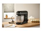 Black stand mixer with stainless steel bowl, baking ingredients, and accessories on a wooden counter.
