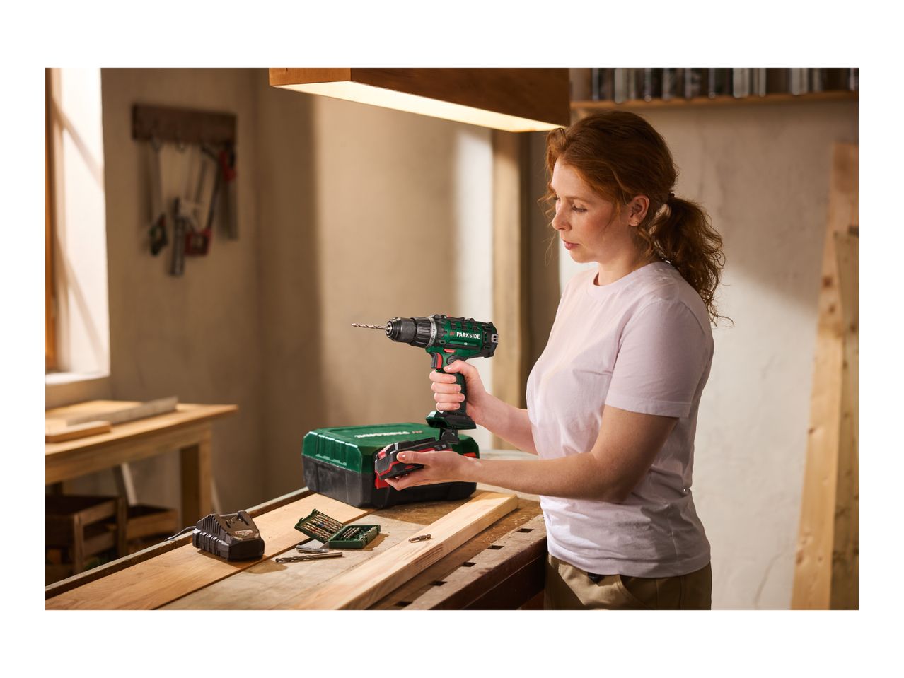 Woman holding a cordless drill, with a battery charger and drill bits on a wooden workbench.