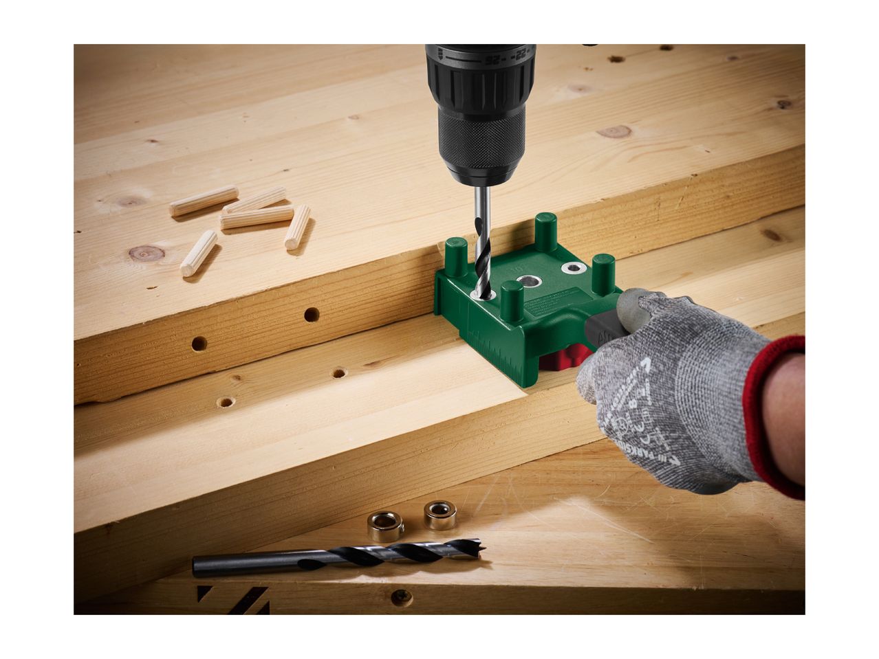 Drilling holes in wood with a drill and a green dowel jig, with wooden dowels nearby.
