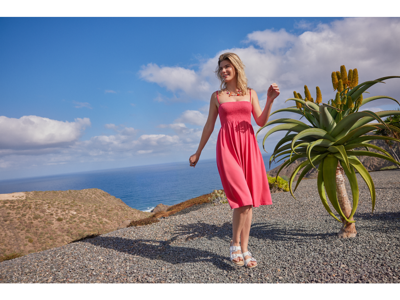 Femeie în rochie roz ESMARA, sandale albe, cu marea și un aloe în fundal.