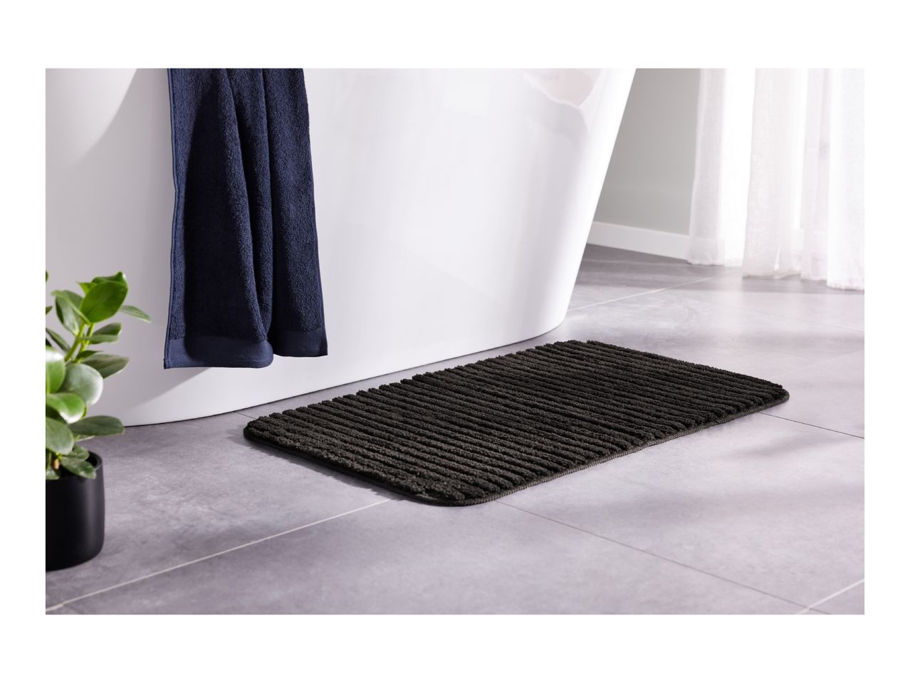 Dark grey bath mat on a tiled floor next to a white bathtub with a navy towel.