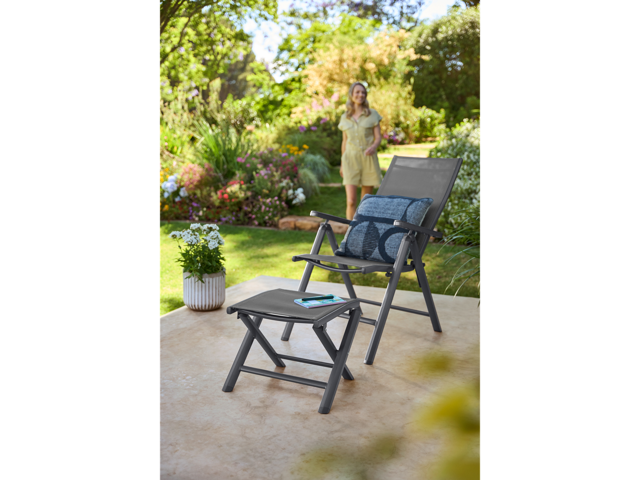 Livarno aluminijumska sklopiva stolica i oslonac za noge na terasi, žena u bašti u pozadini.