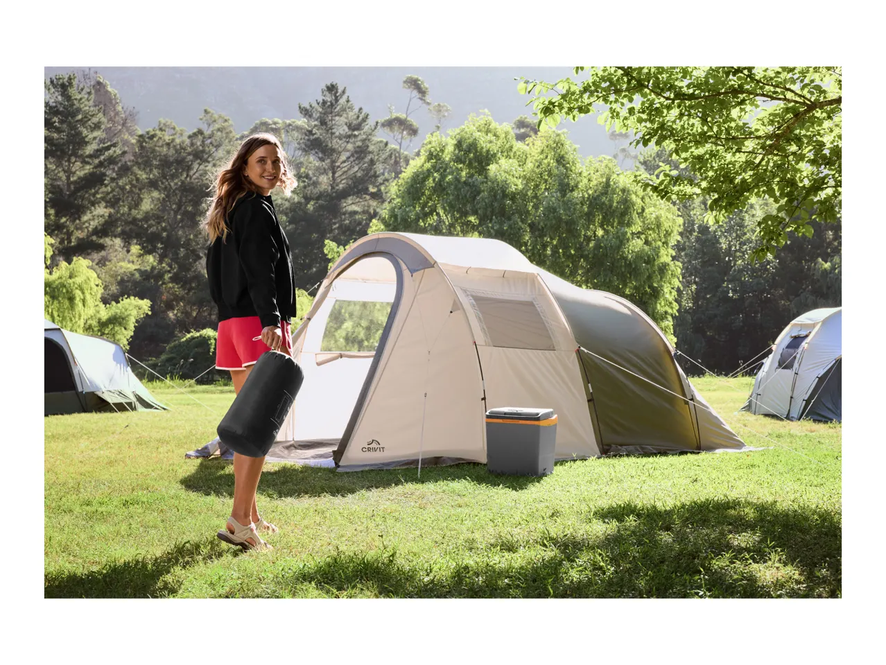 Woman holding a sleeping bag next to a camping tent and a cooler in a grassy field.
