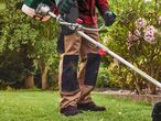 Person in workwear using a gas-powered brush cutter in a garden.