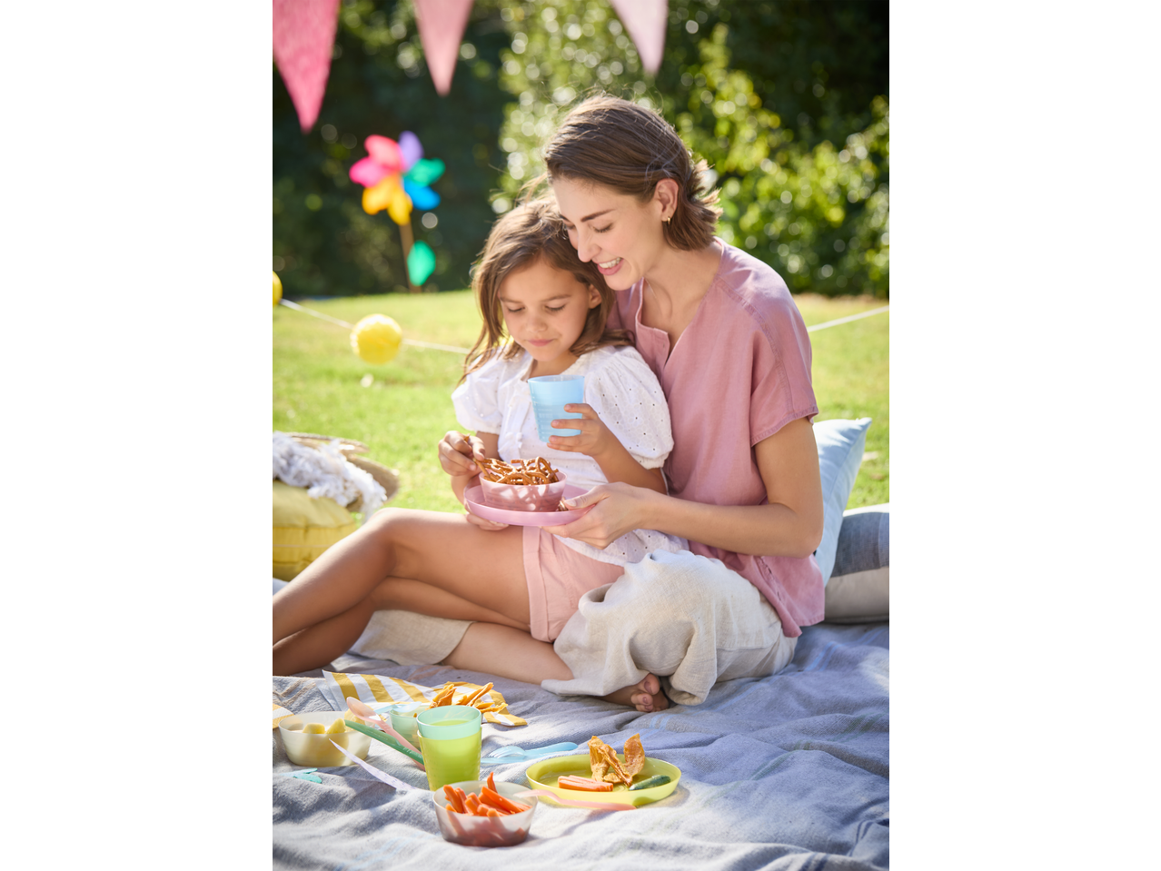 Mother and daughter enjoying a picnic with colorful plastic cutlery and bowls on a blanket.