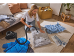 Woman using a vacuum cleaner to vacuum pack clothes into storage bags on a rug.