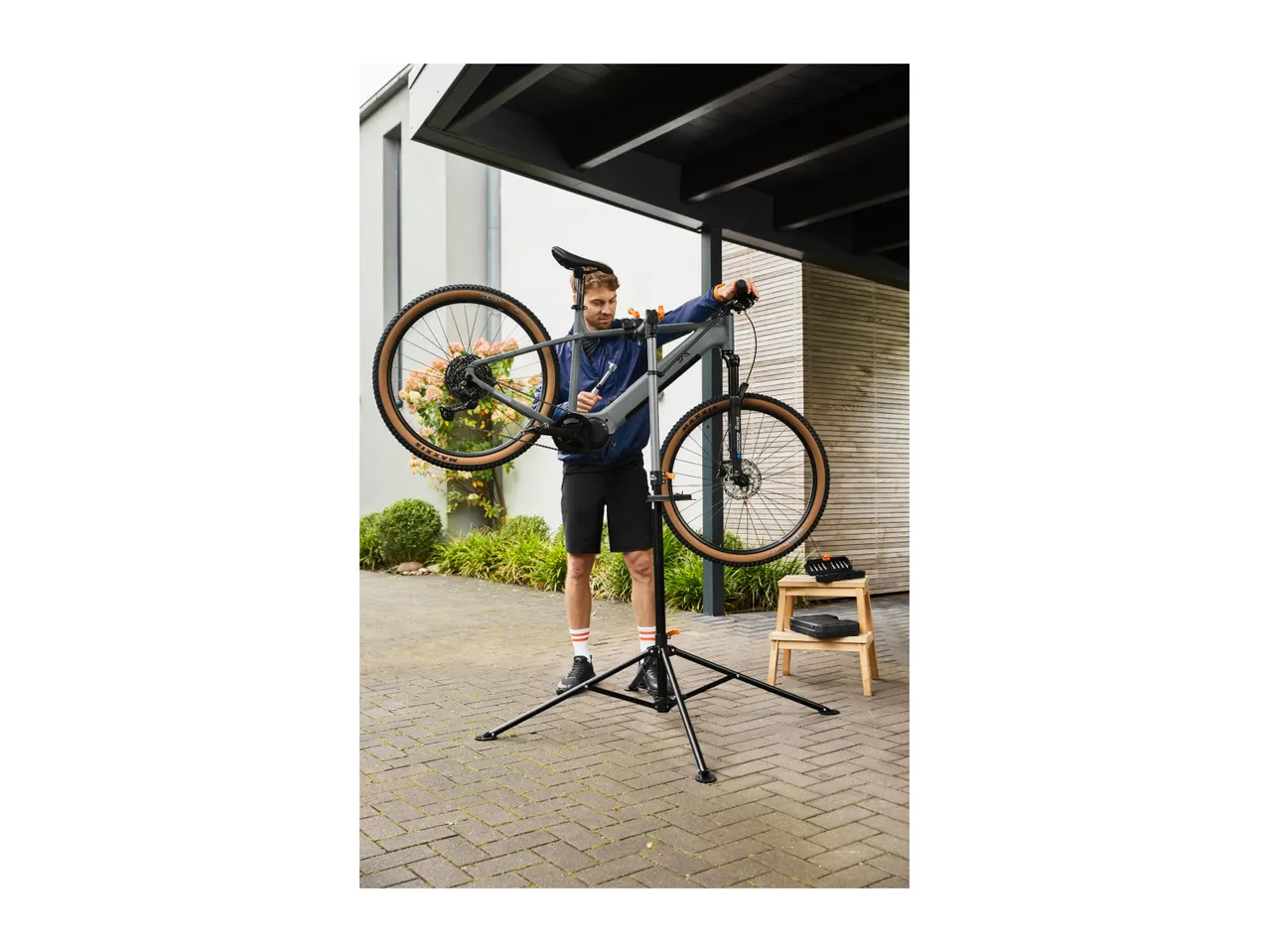 Man repairing a bicycle on a repair stand in a driveway, with tools on a stool nearby.
