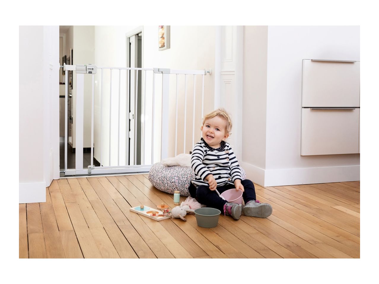 Happy baby sitting on a wooden floor next to a white Safety 1st baby gate.