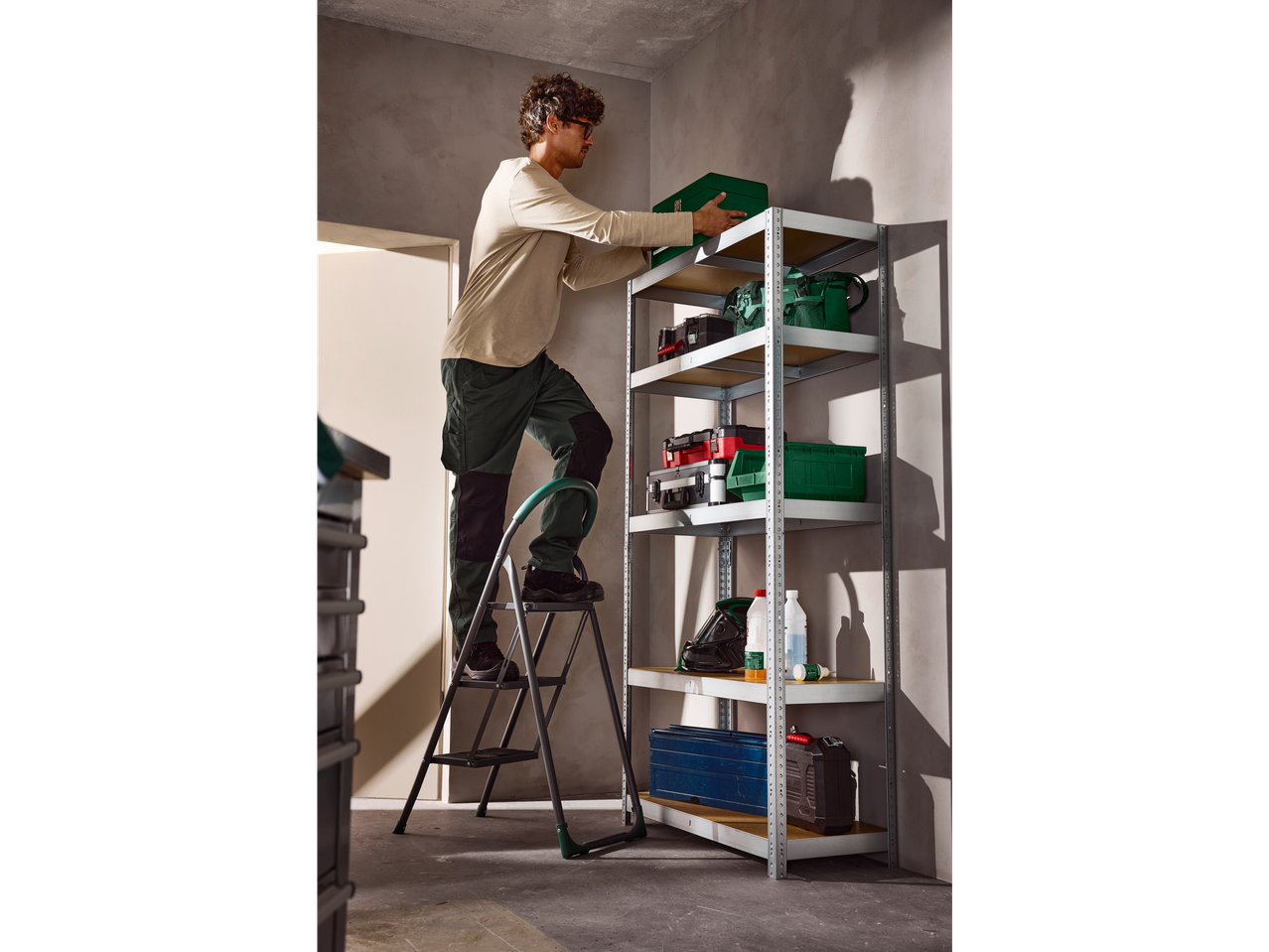 Άνδρας σε σκαμπό Parkside® 3 σκαλοπατιών, τακτοποιεί εργαλεία σε ράφι.