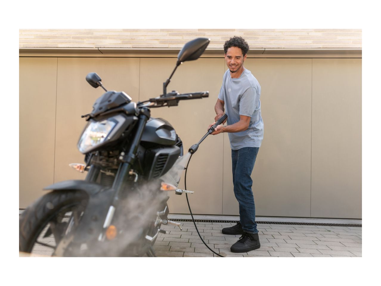 Man pressure washing a motorcycle in front of a modern building.