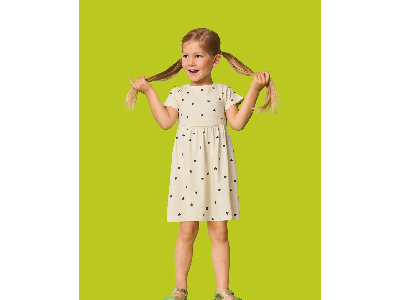 A young girl in a cream dress with black hearts, holding her pigtails, on a lime green background.