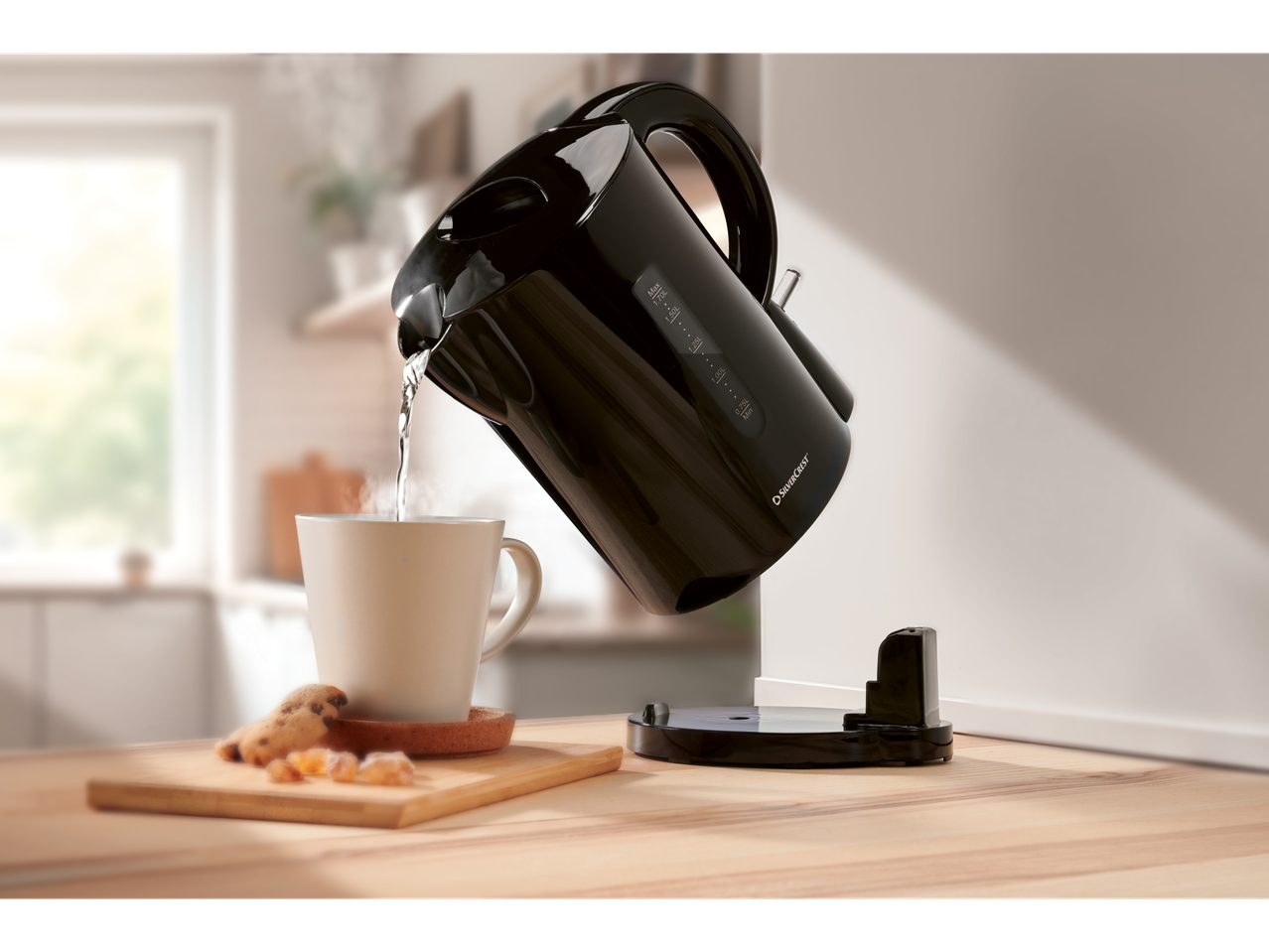 Black SilverCrest kettle pouring hot water into a mug on a wooden table.