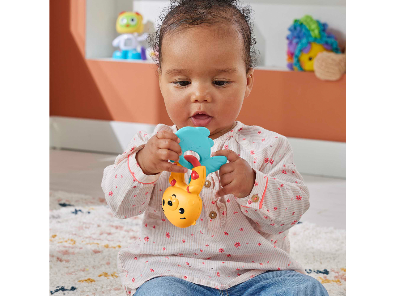 Fisher-Price baby holding a yellow and blue Rattle, sitting on a patterned rug.