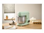 A mint green stand mixer with a stainless steel bowl on a wooden counter with baking ingredients.