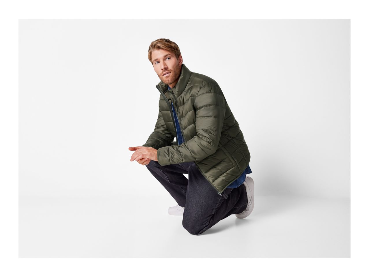 Man in an olive green puffer jacket, blue denim shirt, and dark jeans, crouching.