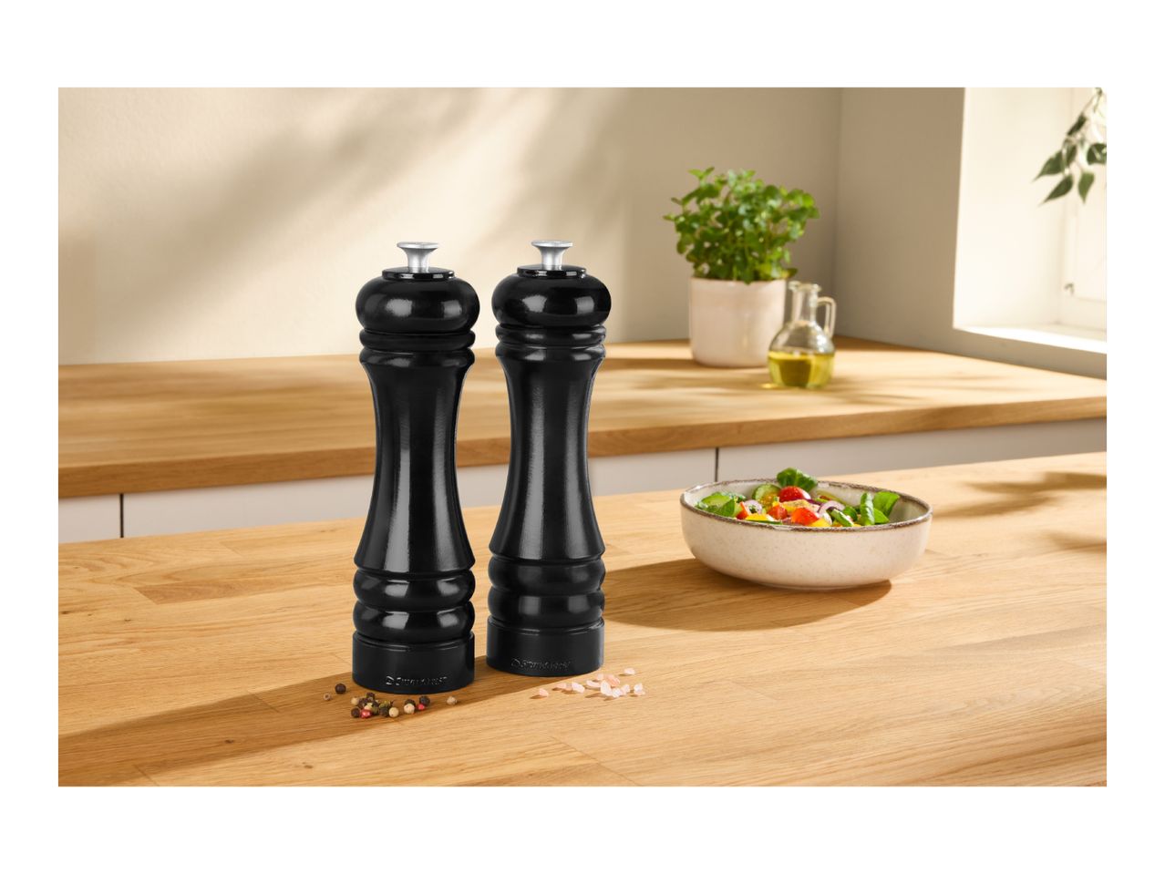 Two black pepper and salt mills on a wooden kitchen counter with a salad bowl.