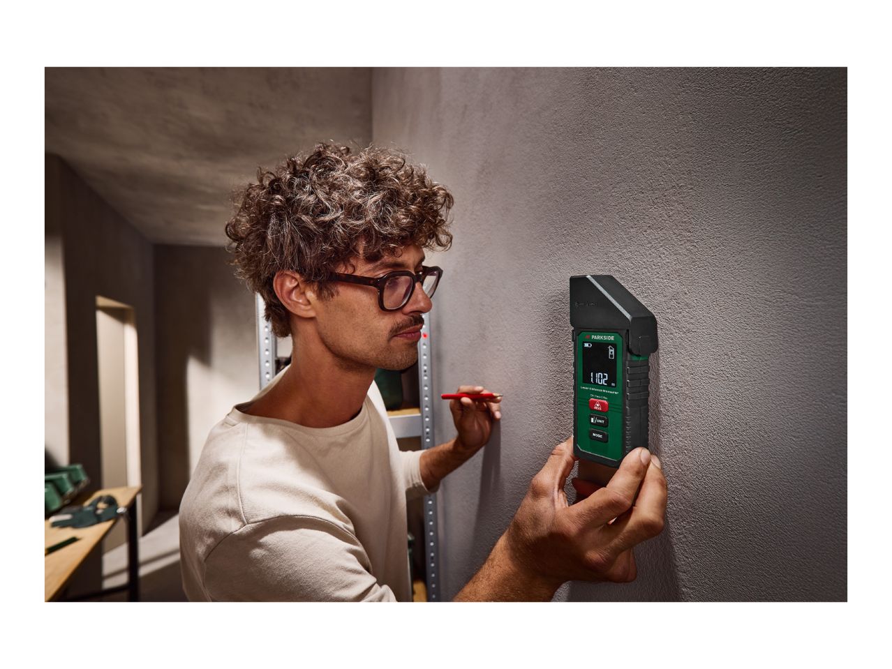 Man using a laser distance measurer on a wall, holding a red pencil.