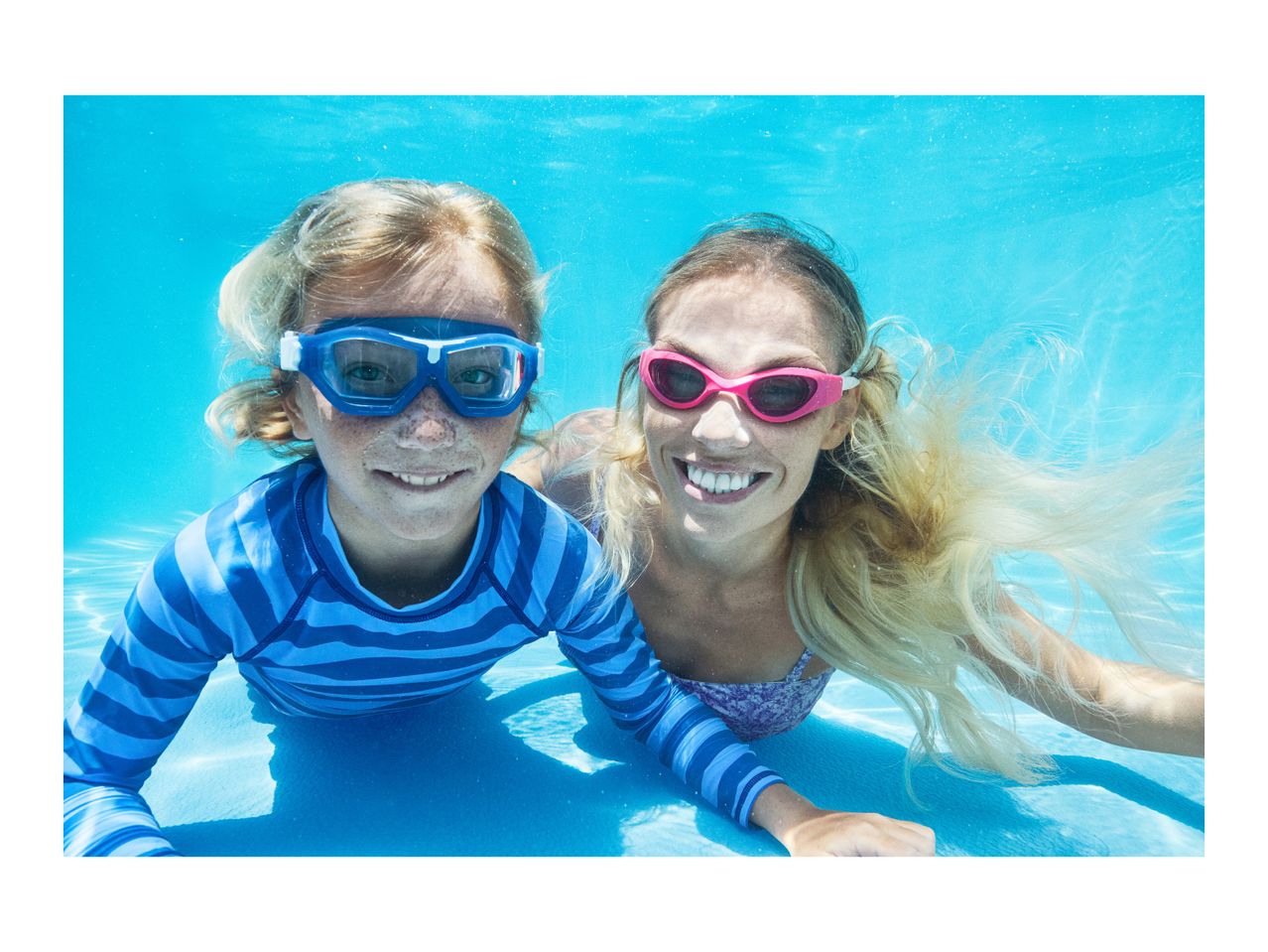 Two people in swim goggles and swimwear underwater, smiling at the camera.
