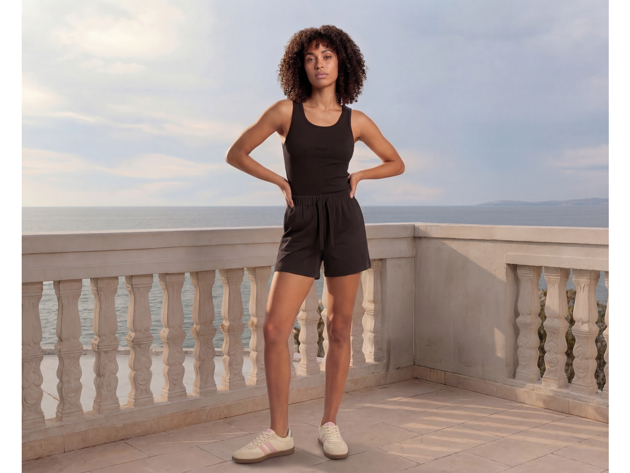Mulher de top preto e calções numa varanda com vista para o mar.