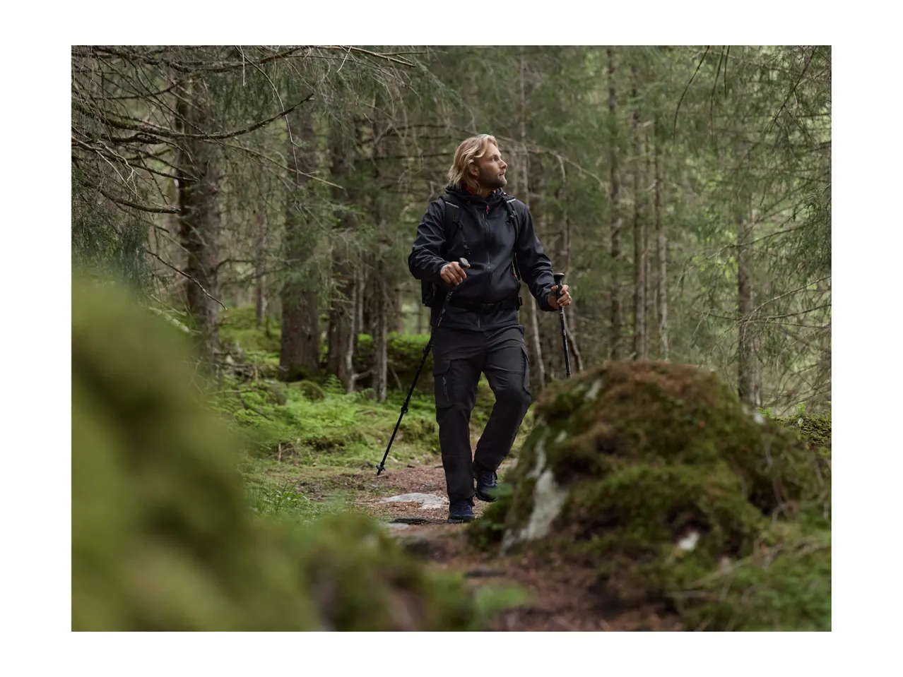 Man hiking in a forest, wearing a black jacket, cargo pants, and using trekking poles.