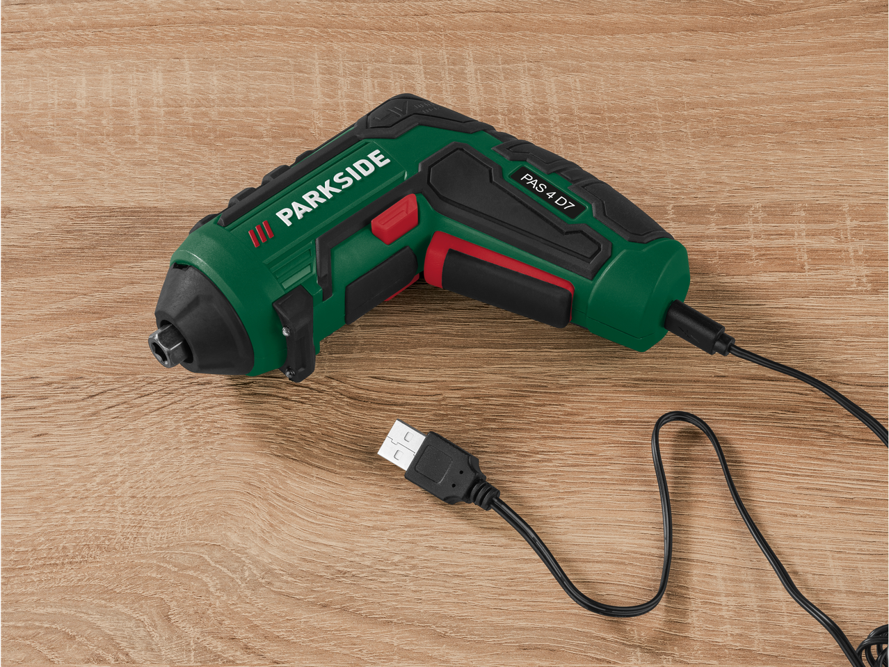 A Parkside PAS 4 D7 cordless screwdriver with USB charging cable lies on a wood surface.