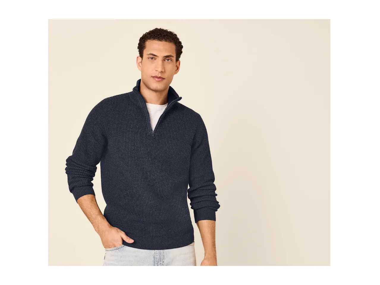 A man wearing a dark blue ribbed half-zip sweater over a white t-shirt and light jeans.
