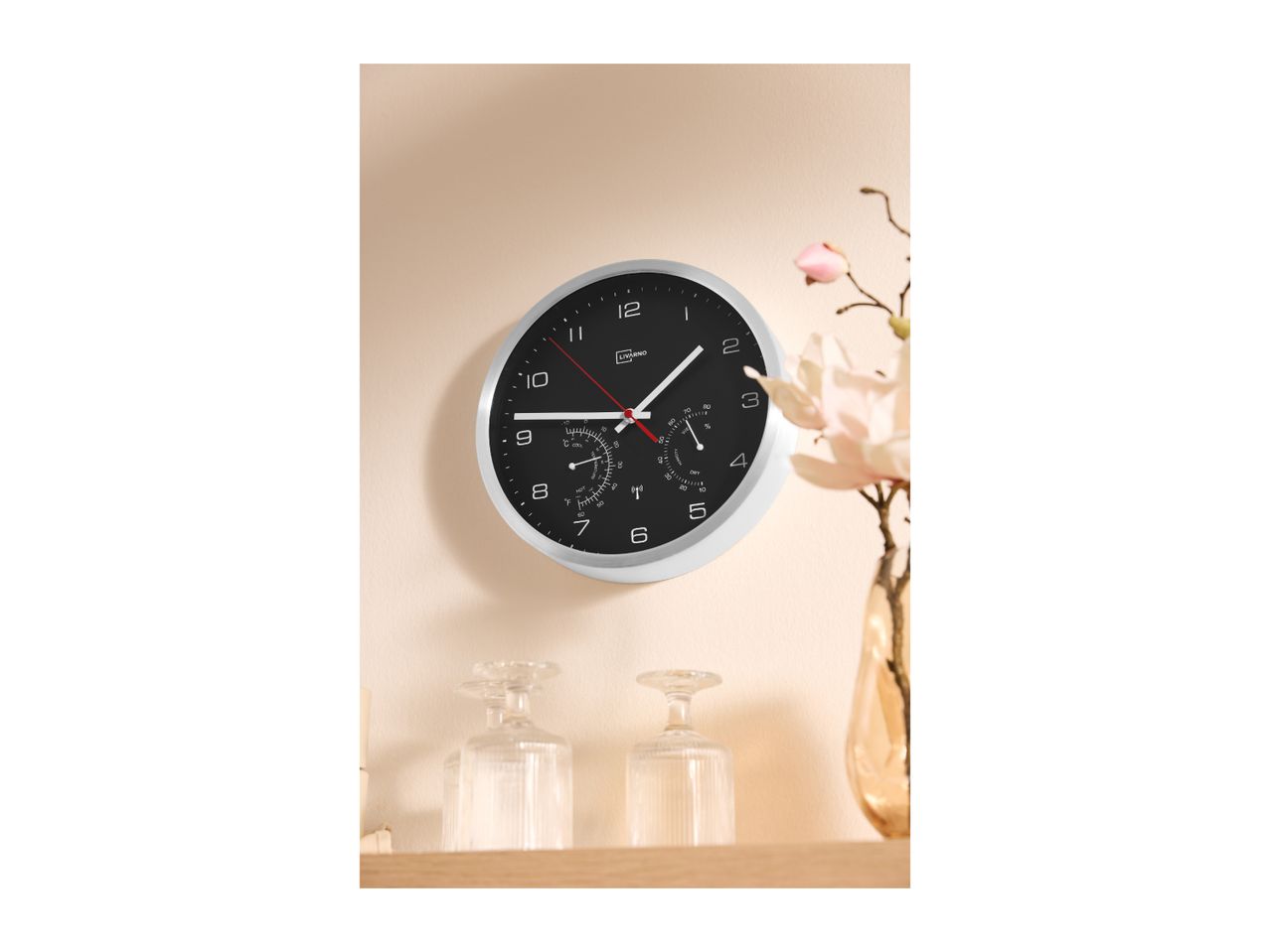 A black and white wall clock with temperature and humidity displays, hanging above two clear glasses and a vase with flowers.