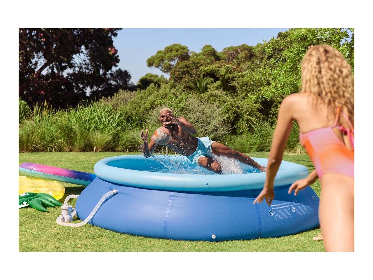 Man jumping into an inflatable pool with a ball, woman watching in a sunny garden.