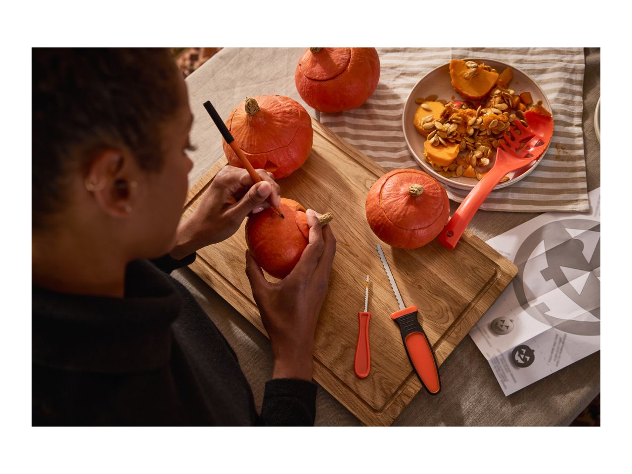 A person carving a small pumpkin on a wooden board with carving tools nearby.