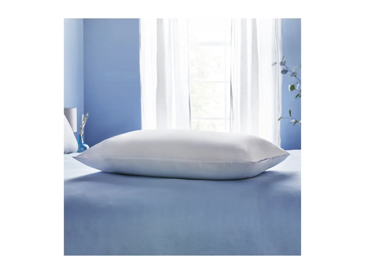 A white Silentnight pillow rests on a blue bedspread in a bedroom with a window.