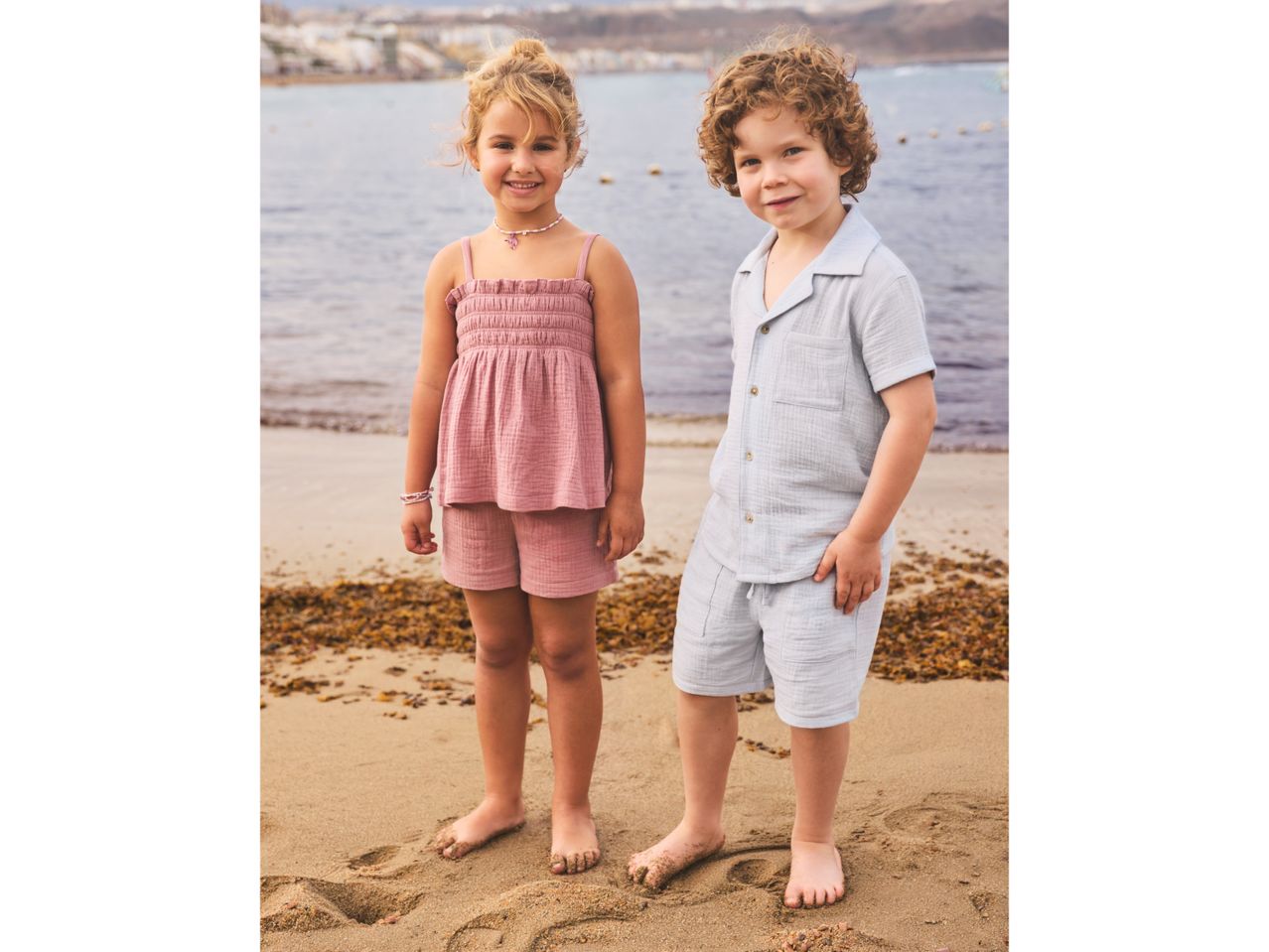 Two children in summer outfits on a beach, a girl in a pink top and shorts, a boy in a light blue shirt and shorts.