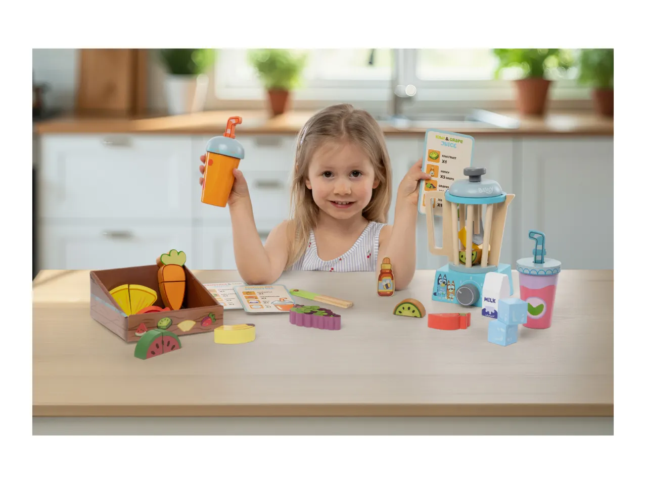 A young girl playing with a toy blender set, holding a toy cup and a recipe card for kiwi and grape juice.