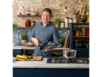 Jamie Oliver ține o tigaie Tefal într-o bucătărie cu o oală pe aragaz și legume pe blat.