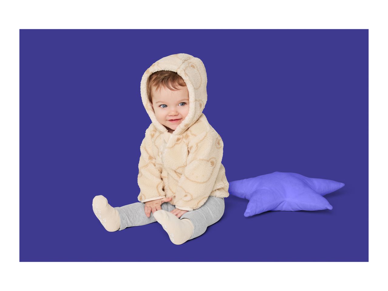 Baby in a hooded fleece jacket and grey leggings, sitting next to a purple star pillow.