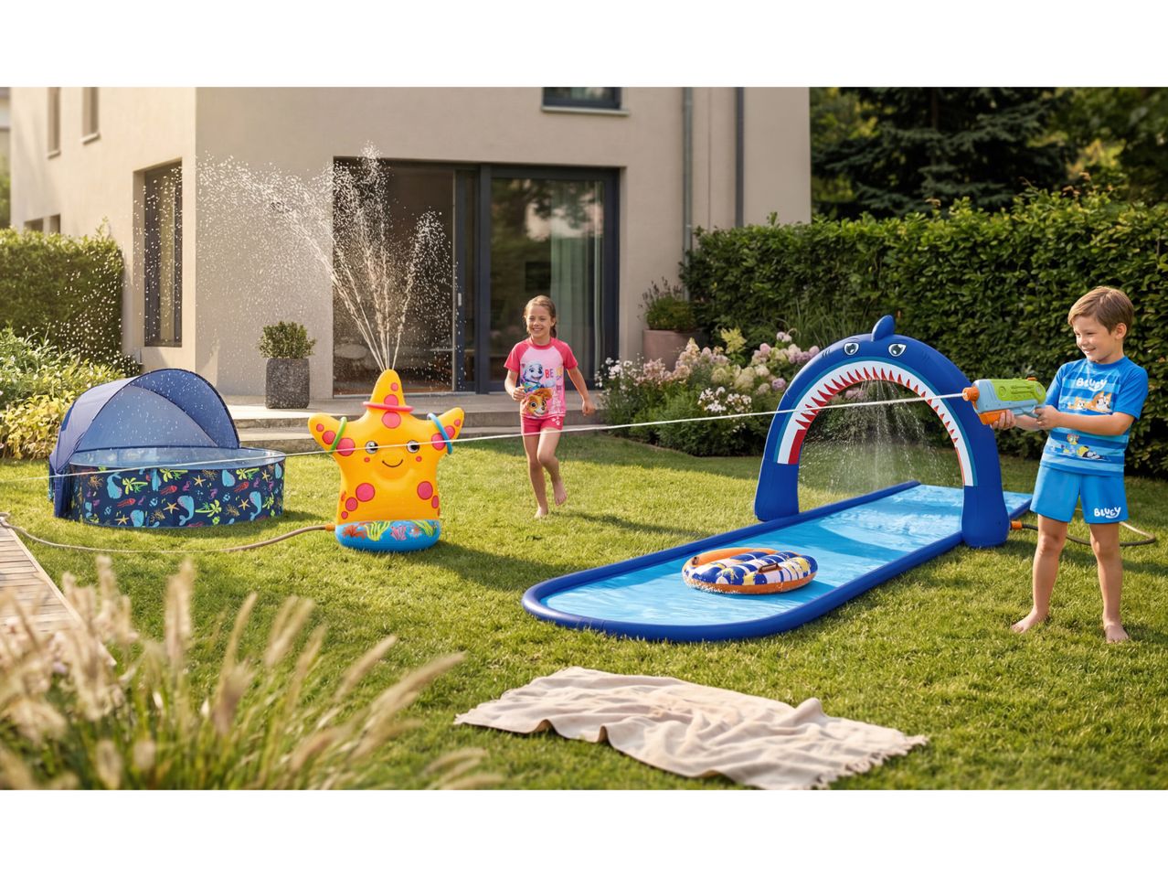 Children playing in a garden with a starfish sprinkler, a shark water slide, and a paddling pool.