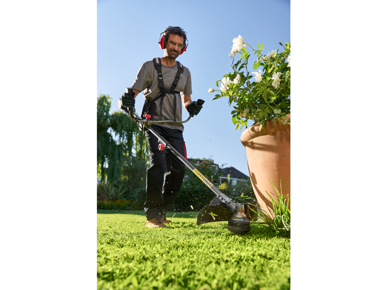 Man using a grass trimmer in a garden, wearing ear protection and safety glasses.