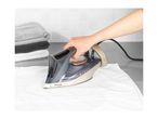 A person ironing a white shirt with a steam iron, with folded towels in the background.