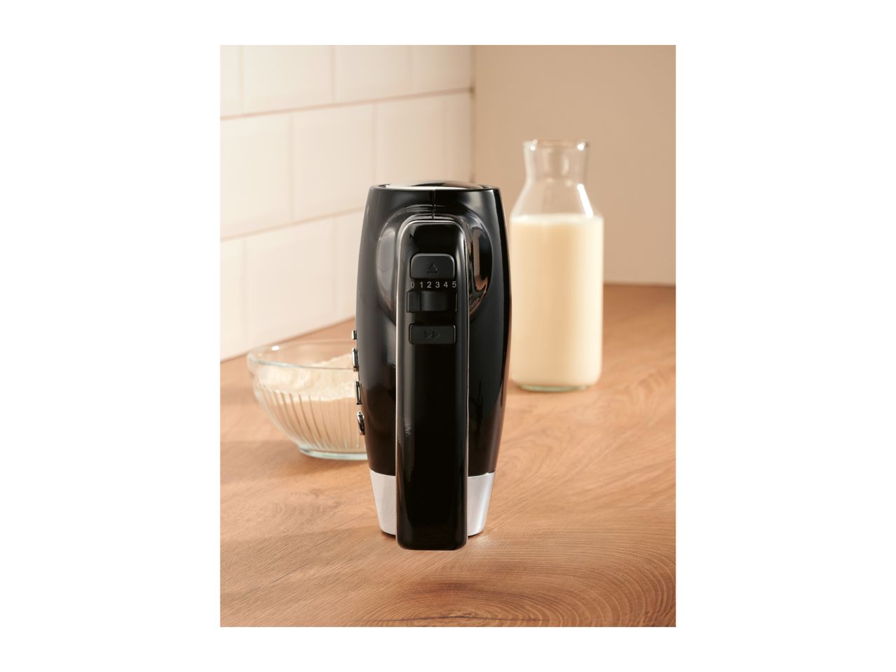 Black hand mixer standing on a wooden countertop next to a bowl of flour and milk