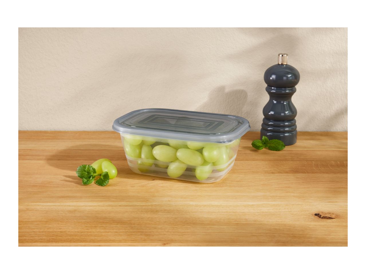 A clear plastic container filled with green grapes on a wooden surface next to a dark gray pepper mill.