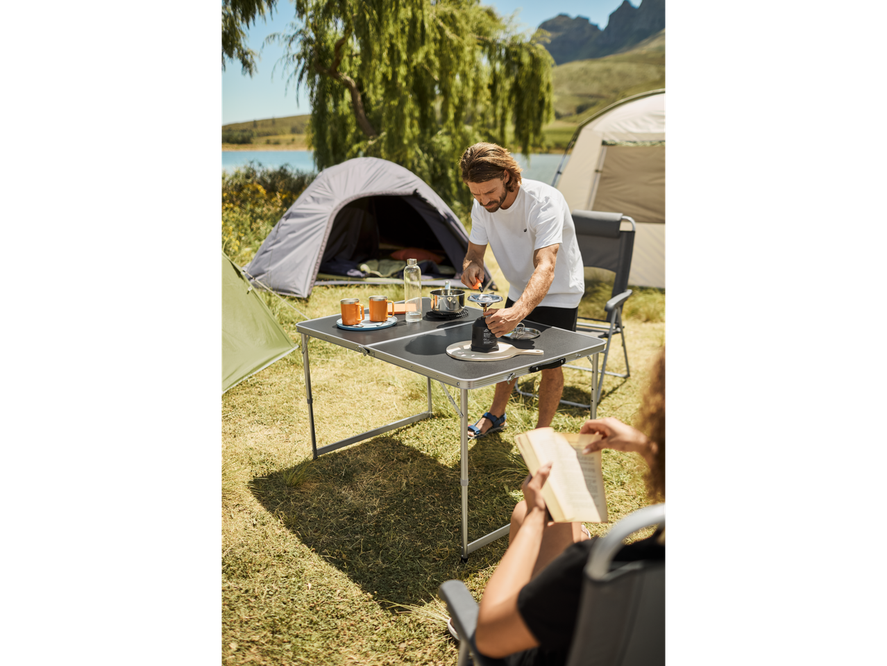 A couple enjoys camping, preparing coffee on a portable camping stove at a foldable table.