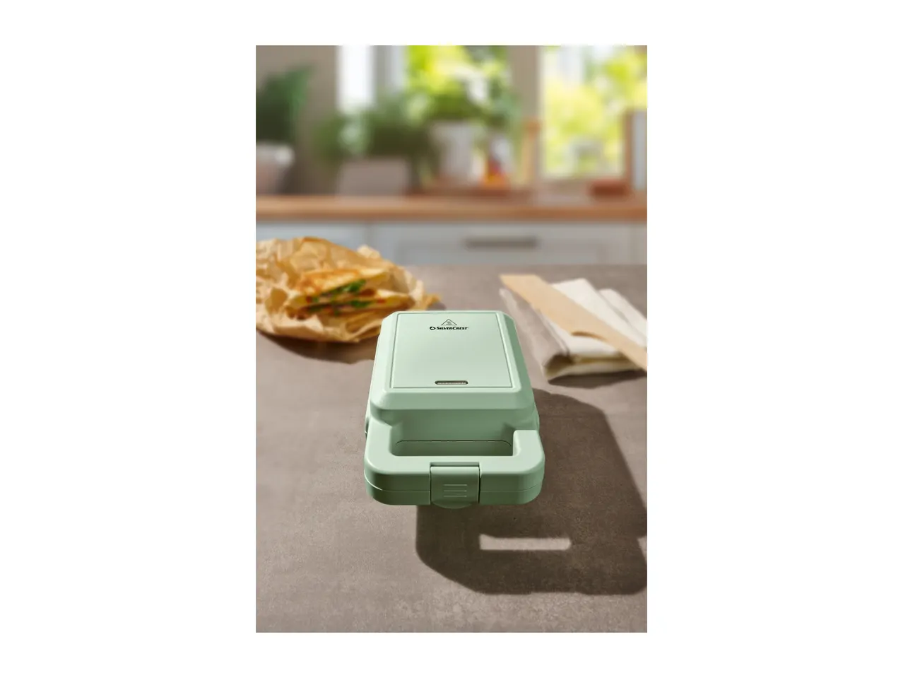 A light green SilverCrest sandwich maker on a kitchen counter with a sandwich in the background.