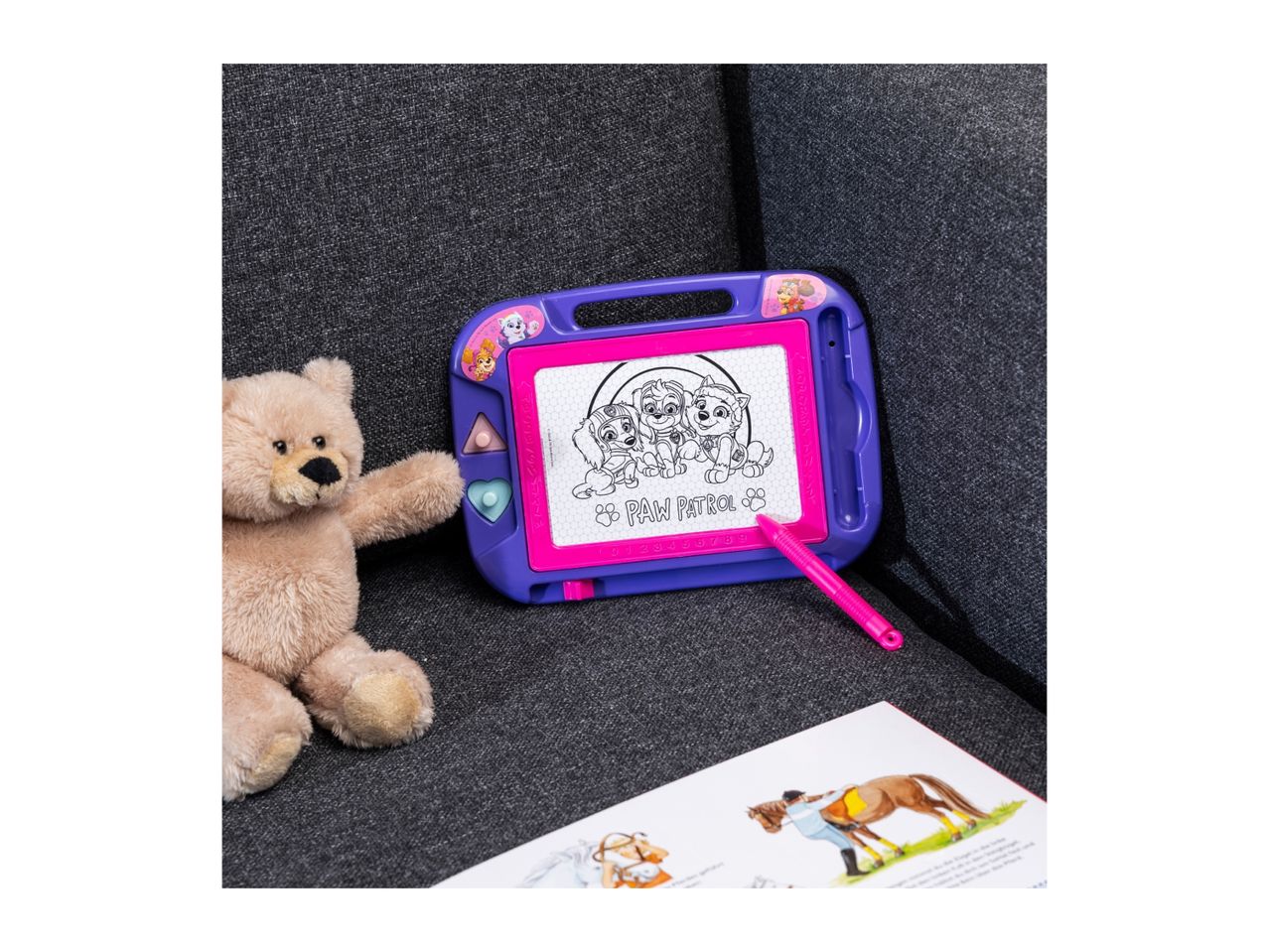A Paw Patrol drawing board with a teddy bear and a book on a gray couch.