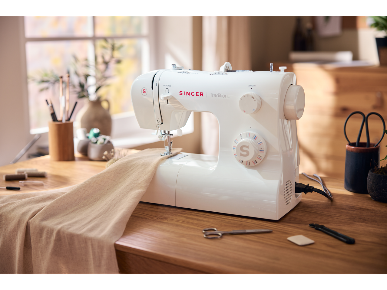 Siuvimo mašina Singer Tradition ant medinio stalo su audiniu ir siuvimo priedais.