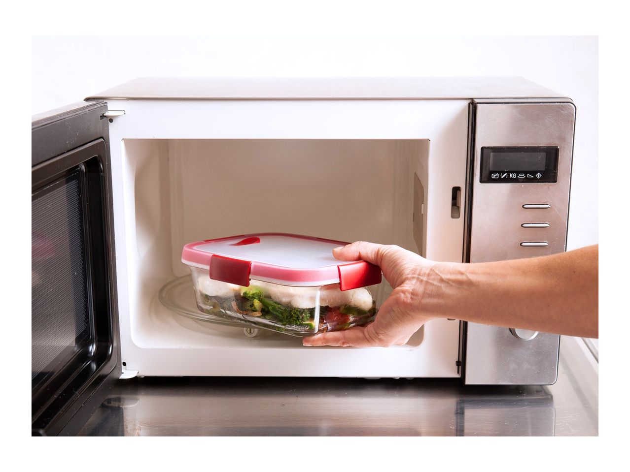 Hand placing a glass food container with vegetables and fish into a microwave.