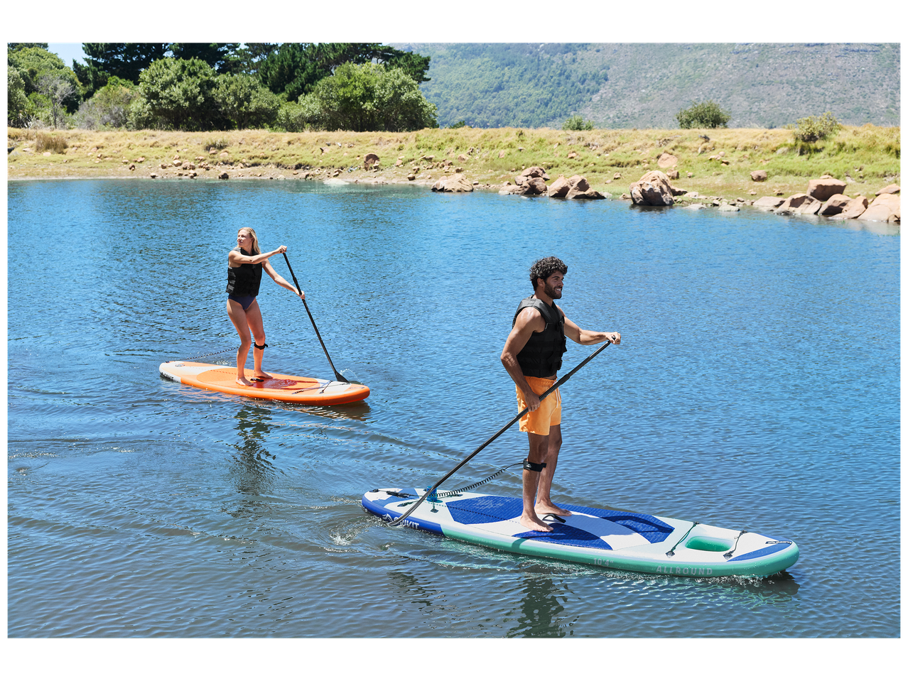 Deux personnes font du paddleboard sur un lac, avec des montagnes et des arbres en arrière-plan.
