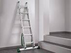 Többcélú alumínium létra zöld lábakkal és hevederekkel, falnak támasztva egy lépcső mellett.